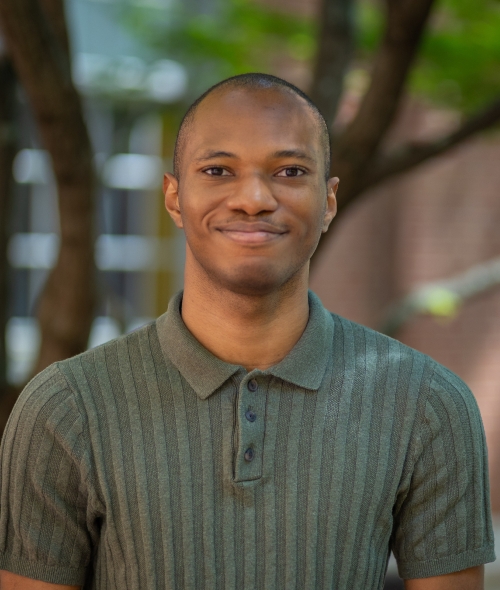 Portrait of Brillon Joseph wearing a green short-sleeved polo shirt, smiling outdoors in front of a brick building and trees.