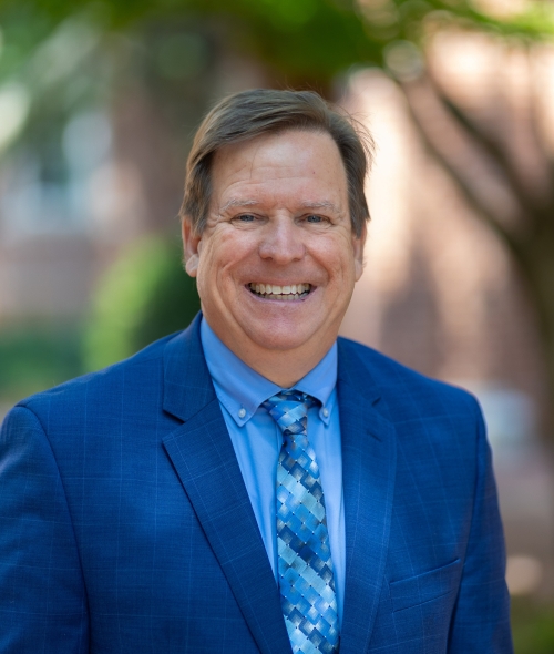 Hugh Lee smiles broadly in a professional headshot, wearing a royal blue suit jacket, light blue dress shirt, and a blue patterned tie against a softly blurred outdoor campus background.
