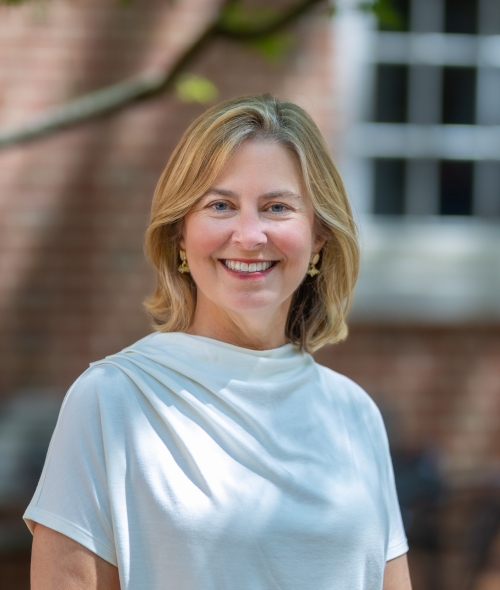 Portrait of Jane Mangan smiling outdoors while wearing a white short-sleeved blouse against a soft-focus brick building background.