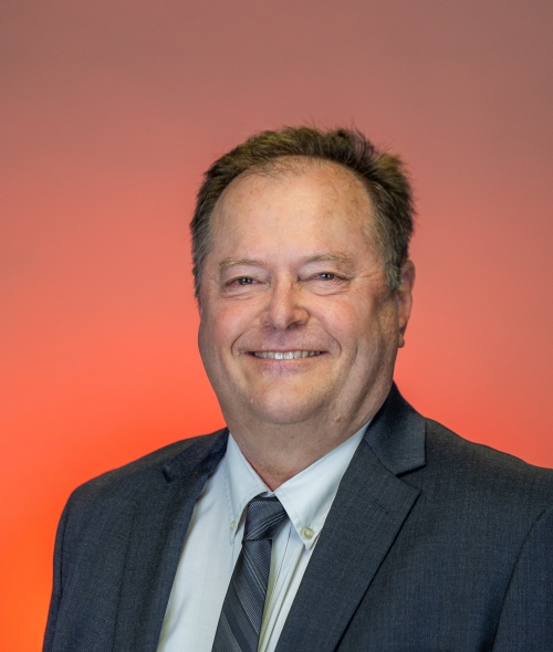 Kenneth Menkhaus smiles in a professional headshot, wearing a dark gray suit jacket, a light dress shirt, and a gray patterned tie against an orange and neutral gradient background.