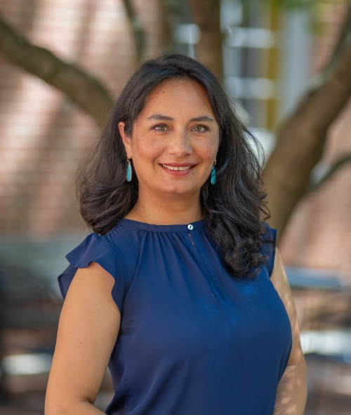 Portrait of Marcela Bacon wearing a dark blue short-sleeved top and turquoise drop earrings, smiling outdoors.