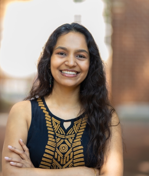 A smiling young woman with long, dark, wavy hair and arms crossed in a sunlit outdoor courtyard