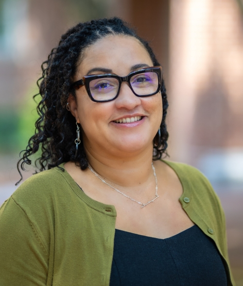 Trasheil Sinnett smiles in a headshot, wearing thick black-framed glasses, dark braided hair, and an olive green cardigan over a black top, with a softly blurred background.