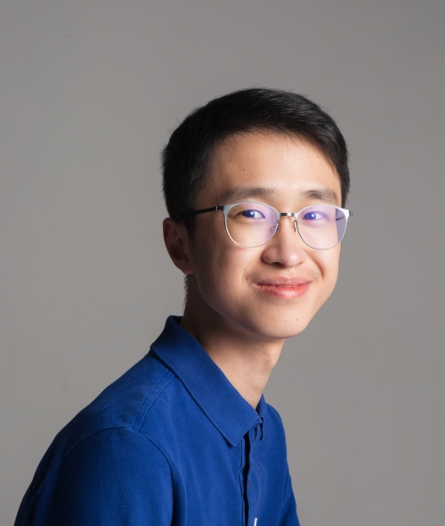 A male student with glasses and a blue button down smiles softly at the camera with head turned