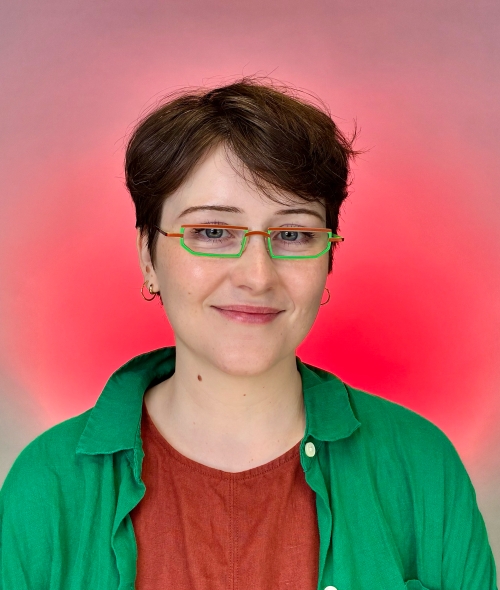 Headshot of a person with short brown hair smiling, wearing colorful orange and green glasses, a rust-red shirt, and a bright green jacket against a pink gradient background.