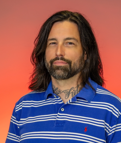 A man with shoulder-length dark brown hair, a full beard, and neck tattoos looking toward the camera against a warm red gradient background. He is wearing a blue polo shirt with horizontal white stripes and a small red logo on the chest.