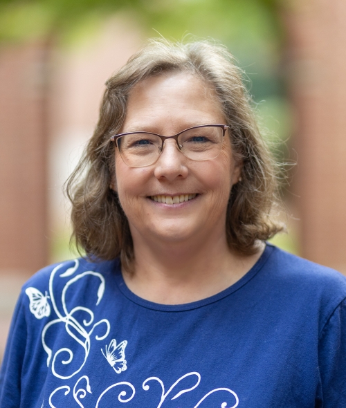 A smiling woman with light brown hair, glasses, a blue embroidered blouse stands in an outdoor courtyard