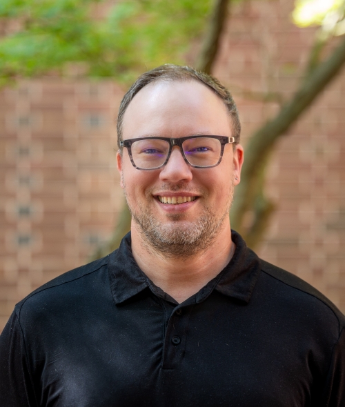 A man with glasses, short light facial hair, and a black polo smiles in an outdoor courtyard