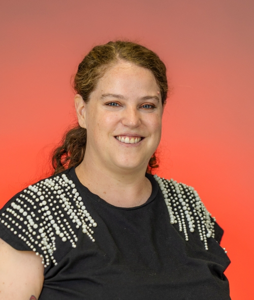 A person with a dark, beaded shirt, red curly hair, hazel eyes and light skin on a red background