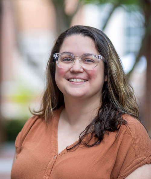 A smiling woman with wavy brown hair, light skin with freckles, and clear glasses stands in an outdoor courtyard