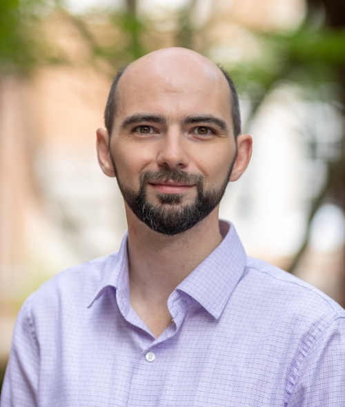 A light-skinned man, outdoors, with a purple button-down, no hair, and brown facial hair