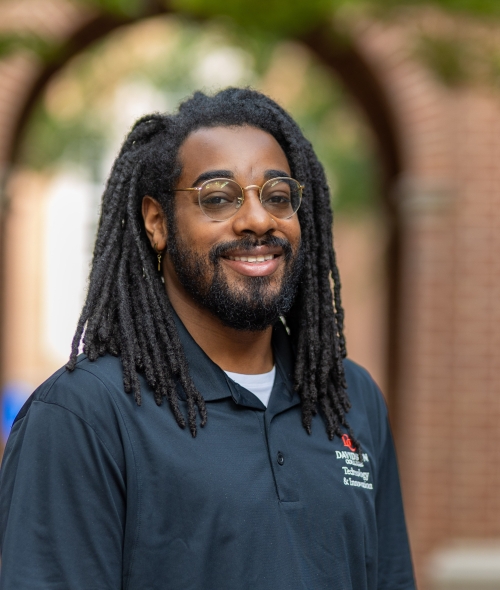 A smiling man with shoulder-length braids, wire-rimmed glasses, and a full beard wears a black "Davidson College" polo shirt, standing in an outdoor courtyard