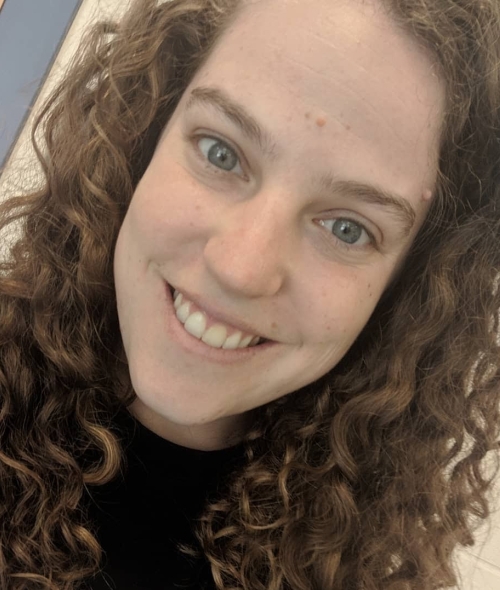 A person with long, curly brown hair and hazel eyes smiling directly at the camera.