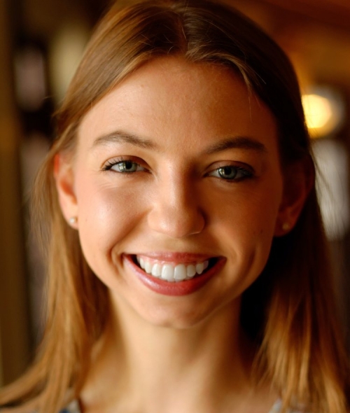 Close-up portrait of a smiling young woman with light brown hair, wearing a pattern-printed top.