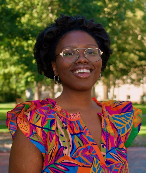 A Black woman wearing wire-rimmed glasses, silver hoop earrings, and a brightly colored tropical-print top smiles outdoors with lush green trees in the background.