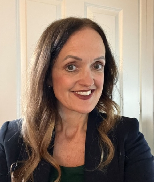 A woman with long, wavy brown hair with light ends smiles while wearing a dark blazer over a green top in front of a white door.