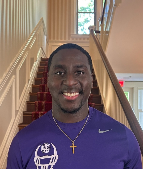Portrait of a smiling young Black man wearing a purple Nike t-shirt with a football helmet graphic, a gold cross necklace, standing in front of a grand wooden staircase with a red patterned runner carpet.