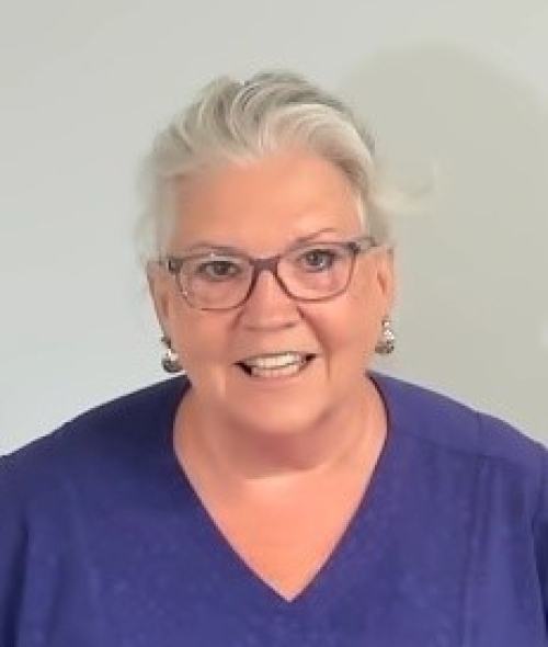Headshot of a smiling woman with styled white hair, wearing dark glasses, silver drop earrings, and a bright blue v-neck shirt against a neutral background.
