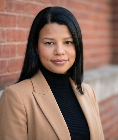 A smiling woman with shoulder-length dark hair wearing a camel blazer and a black turtleneck standing in front of a red brick wall