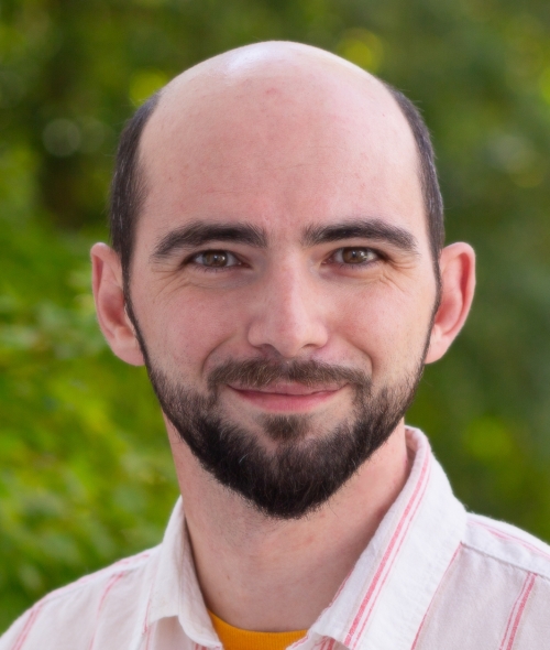 A smiling, light-skinned man with a bald head, dark brown eyes, and a full dark beard against a blurred green background.