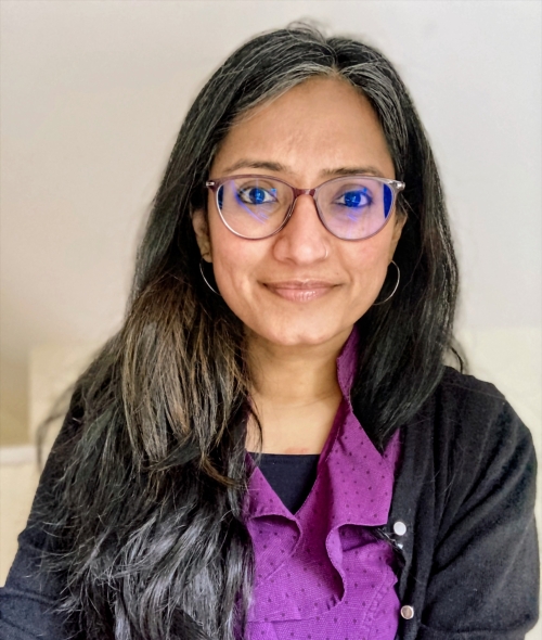 A headshot of a woman with long grey-streaked dark hair, wearing glasses, thin hoop earrings, and a purple patterned top paired with a black cardigan.