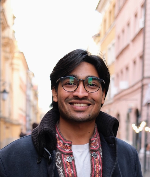 A male student wtih a navy blue jacket and colorful shirt, glasses, standing on a city street