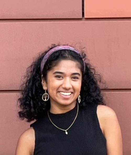 A woman with dark curly, shoulder-length hair in a purple headband stands in front of a red terra cotta wall