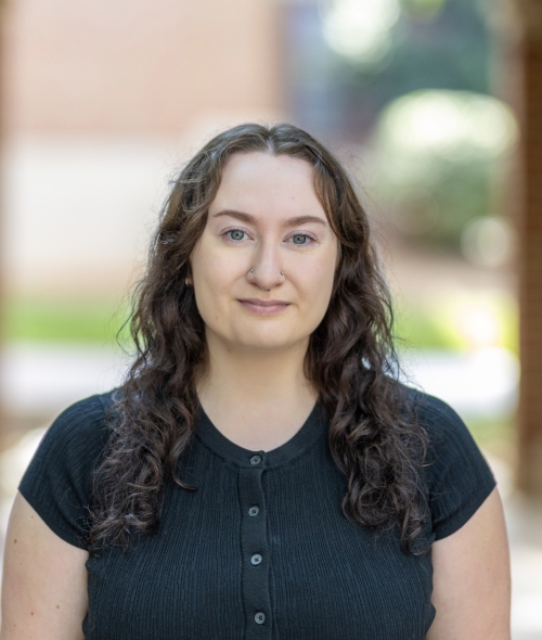 An adult woman in a blue shirt with light skin and curly dark hair stands outdoors in a courtyard