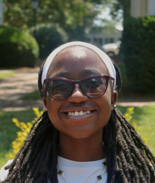 A smiling female student with glasses, white shirt, long dark hair in intricate braids and headband