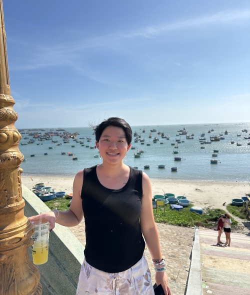 A smiling person with short dark hair and a black tank top holds a cold drink while standing on stairs overlooking a sandy beach dotted with round coracle boats and floating vessels on clear blue ocean water.