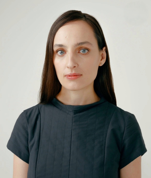 A woman with long dark hair and light eyes looking directly forward, wearing a dark short-sleeved top against a plain light background