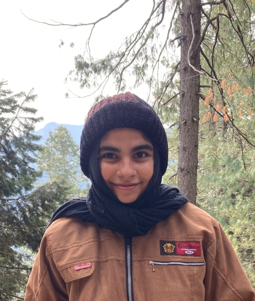 A female student in a forest with a hat and scarf covering her hair, brown canvas jacket, and a mountain view in the background