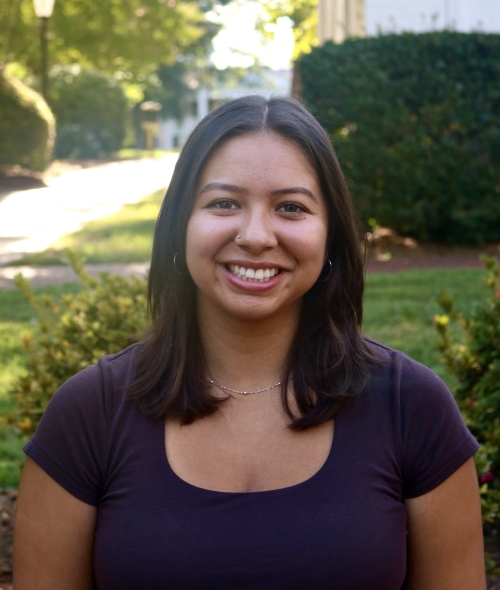 Alex smiling outdoors in a dark scoop-neck top with shoulder-length dark hair