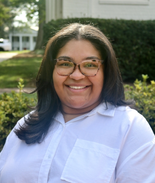 Mariah smiling outdoors wearing glasses and a white collared shirt with shoulder-length dark hair.