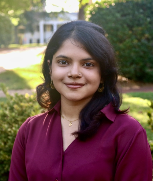 Mishti smiling outdoors in a deep plum-colored collared shirt with shoulder-length dark hair