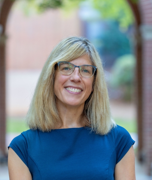 A woman with shoulder-length blonde hair, glasses, and a blue cap-sleeve blouse smiles in an outdoor courtyard