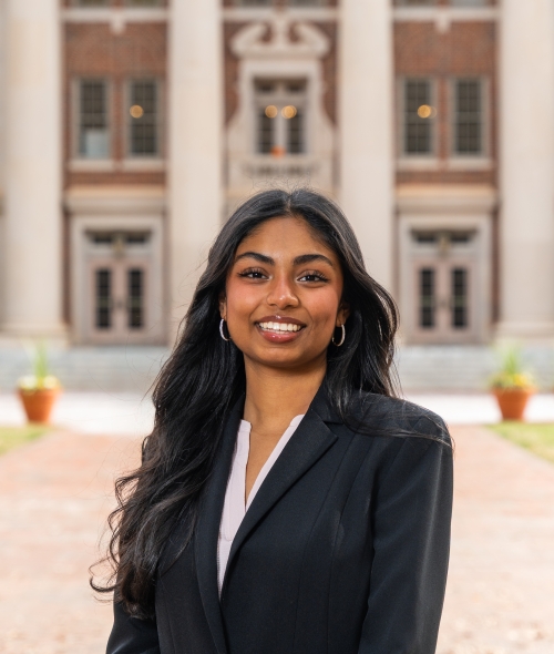 A smiling woman with long dark wavy hair wearing a black blazer over a light shirt standing in front of a brick building.