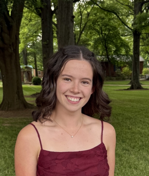 A smiling woman with dark, shoulder-length wavy hair wears a maroon dress and necklace while posing outdoors in front of tall trees