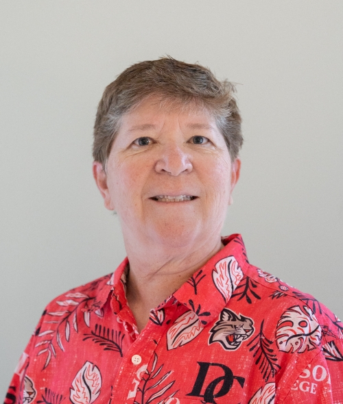 A person with short, brown hair and a smiling expression wears a red patterned collared shirt while posing in front of a grey backdrop
