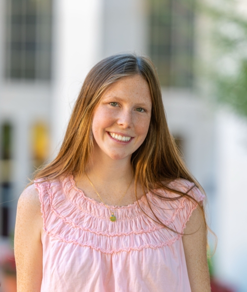 A smiling woman with long straight light brown hair wearing a sleeveless pink top and a gold pendant necklace outdoors