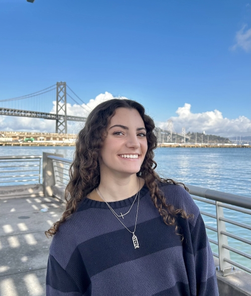 A smiling woman with long curly brown hair wears a dark striped sweater and layered necklaces while posing in front of a bay and bridge under a bright blue sky
