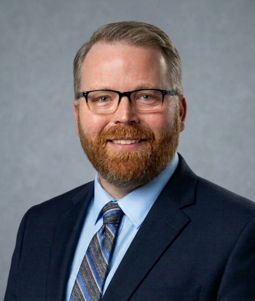 A professional man with short, styled brown hair, glasses, and a neatly trimmed brown and gray beard, wearing a navy blue blazer, light blue dress shirt, and patterned blue tie, smiling against a gray background.