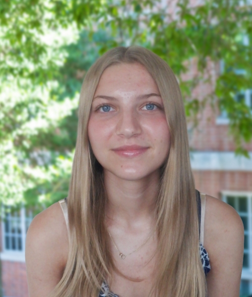 A smiling woman with long, straight blonde hair and light blue eyes wears a simple necklace in front of soft green foliage and a brick building
