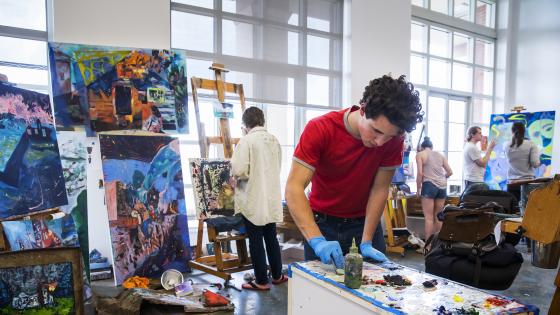 Student mixes paint in foreground while other students paint canvases in background