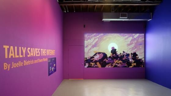 Tally Saves the Internet Exhibit text on wall in purple room, with screen projecting creature on pile of rubble
