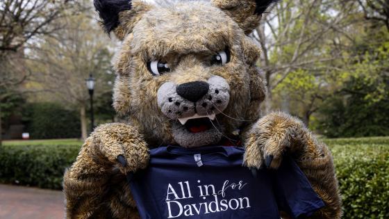 Mascot Lux the Wildcat holds t-shirt that says "All in for Davidson" outside on campus