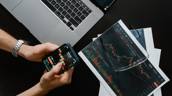 Handings holding phone overlooking laptop and stock projections on piece of paper