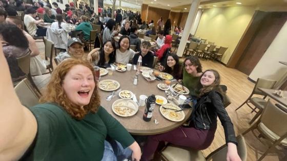 A group of students take a selfie together while sitting around a round dining table in a dining hall