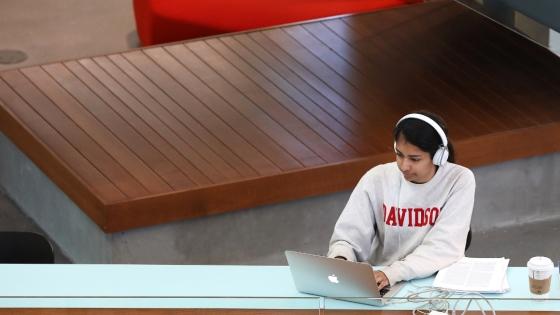 student wearing a Davidson sweatshirt and headphones works on a laptop in a modern academic building