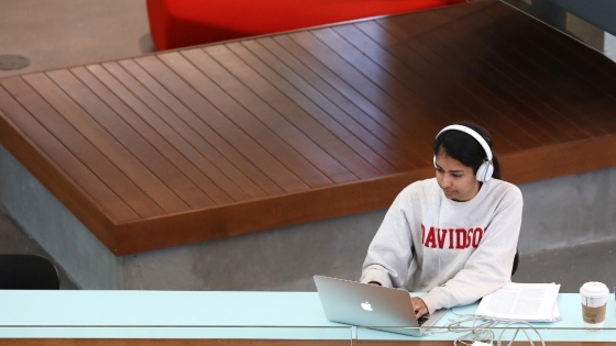 student life paragraph essay student wearing a Davidson sweatshirt and headphones works on a laptop in a modern academic building